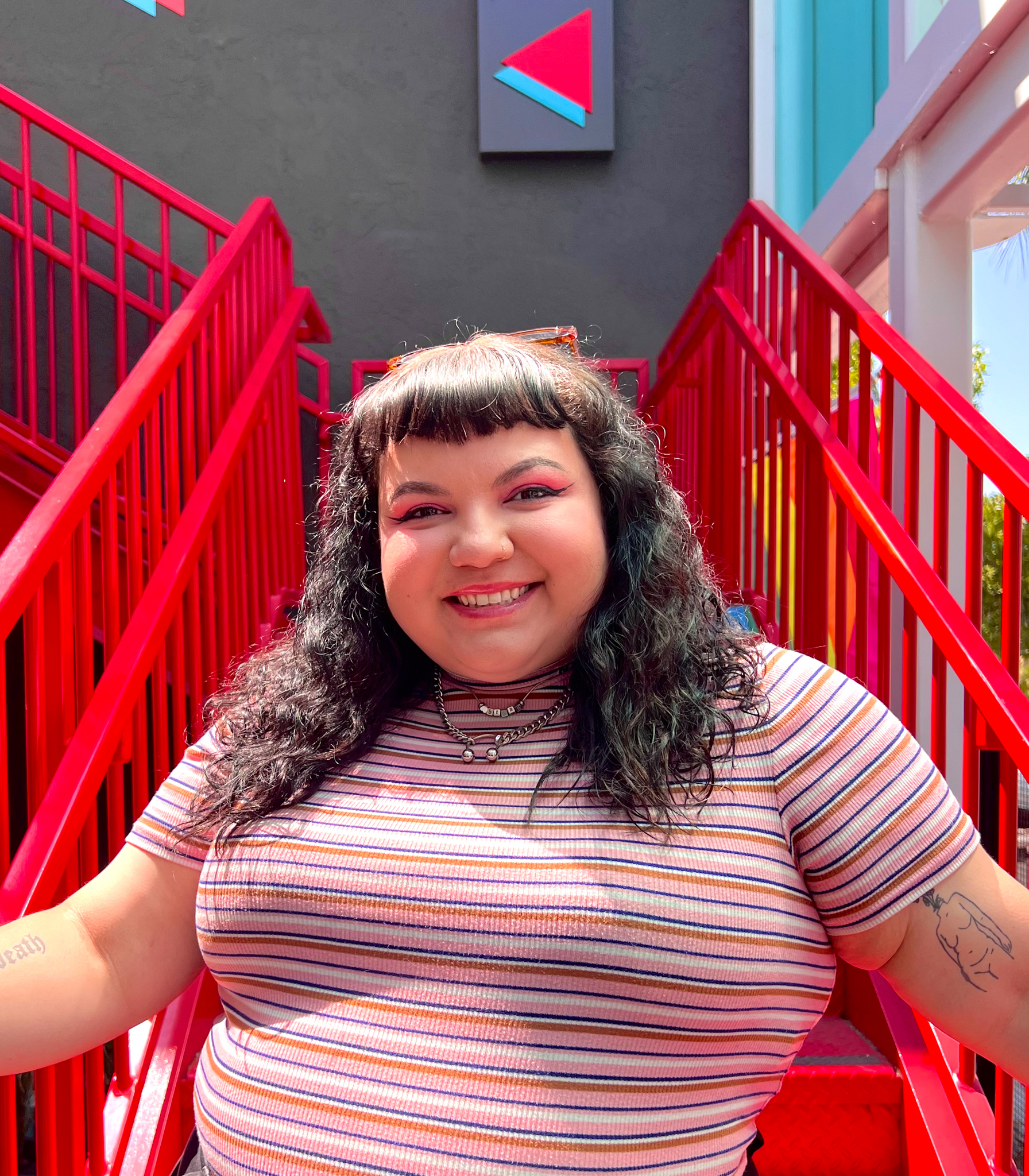 A photo of Natalie Dominguez, Diversionary's Marketing Manager, standing at the bottom of Diversionary's bright red staircase that leads up to the Mainstage. Natalie is wearing a pink striped tops with sunglasses on her head, smiling at the camera.